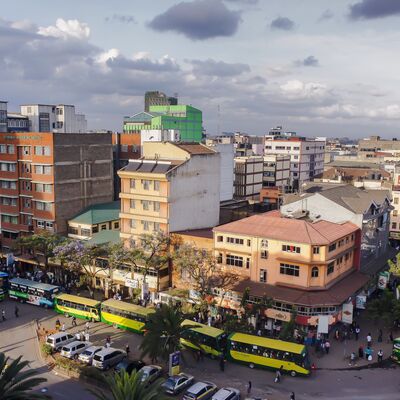 Auf dem Bild ist ein Stra&szlig;enzug in Nairobi abgebildet. Vor dem leicht wolkigen Himmel sind viele aneinander stehende, mehretagige H&auml;user zu sehen. Palmen s&auml;umen die Stra&szlig;e, an dessen Stra&szlig;enrand viele gelb-gr&uuml;ne Kleinbusse stehen.