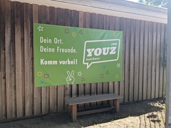 Bild vergr&ouml;&szlig;ern: Am Holzhaus selbst h&auml;ngt das gr&uuml;ne Banner vom Jugendzentrum YOUZ mit der Auschrift: Dein Ort. Deine Freunde. Komm vorbei!