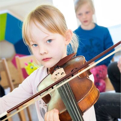Auf dem Foto ist ein weibliche gelesenes Kind zu sehen, das eine Geige h&auml;lt und zum Betrachtenden schaut. Das Kind h&auml;lt die Geige in Spielposition auf der Schulter h&auml;lt sie mit Kinn und linker Hand. Den Geigenbogen hat das Kind zum Spielen auf die Saiten gelegt.
