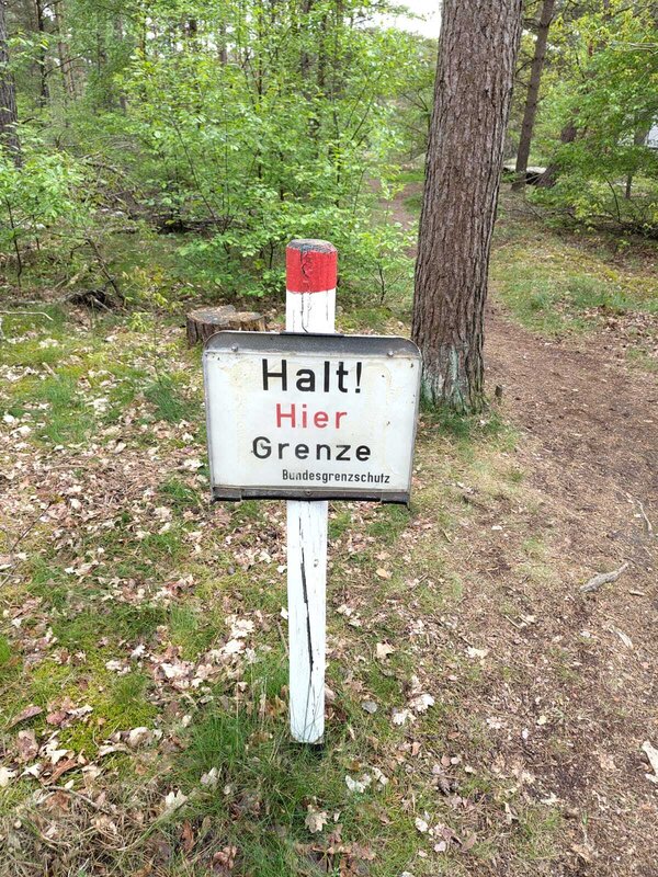 Bild vergr&ouml;&szlig;ern: Auf dem Foto ist ein mitten in einem Wald stehendes Schild zu sehen. Das Bild ist am oberen Ende eines wei&szlig; angestrichenen Holzpfahls zu befestigt. Der Holzpfahl ist am oberen Ende ein kleines St&uuml;ck rot gestrichen. Auf dem Schild mit wei&szlig;em Hintergrund steht geschrieben: "Halt! Hier Grenze Bundesgrenzschutz" Jedes Wort steht dabei in einer eigenen Zeile. Das Wort "Hier" ist in roter Schrift geschrieben, der Rest in schwarzer Schrift.