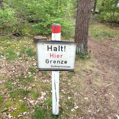 Auf dem Foto ist ein mitten in einem Wald stehendes Schild zu sehen. Das Bild ist am oberen Ende eines wei&szlig; angestrichenen Holzpfahls zu befestigt. Der Holzpfahl ist am oberen Ende ein kleines St&uuml;ck rot gestrichen. Auf dem Schild mit wei&szlig;em Hintergrund steht geschrieben: "Halt! Hier Grenze Bundesgrenzschutz" Jedes Wort steht dabei in einer eigenen Zeile. Das Wort "Hier" ist in roter Schrift geschrieben, der Rest in schwarzer Schrift.