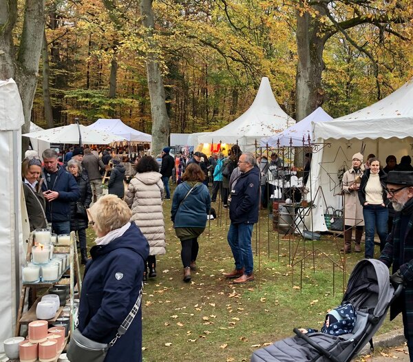 Bild vergr&ouml;&szlig;ern: Auf dem Foto ist ein buntes Marktgeschehen zu sehen. Menschen stehen an St&auml;nden. Die St&auml;nde sind mit wei&szlig;en Pavillons &uuml;berdacht und an und zwischen den St&auml;nden stehen Gartendekorationen, Keramik und vieles mehr. Alles steht auf einer gr&uuml;nen Wiese und ist umgeben von B&auml;umen.
