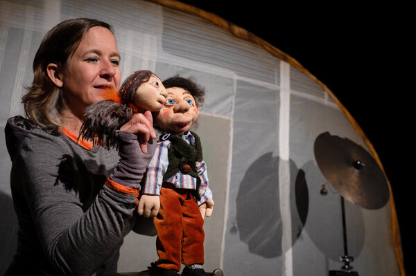 Bild vergr&ouml;&szlig;ern: Auf dem Foto ist auf der linken Seite eine weiblich gelesene Person zu sehen, die zwei Handpuppen in den H&auml;nden h&auml;lt. Eine der Handpuppen ist weiblich gelesen, die andere Handpuppe m&auml;nnlich gelesen. Hinter der puppenspielenden Person befindet sich ein wei&szlig;er Stoff, der aus mehreren Teilen zusammengesetzt wurde. En runder, br&auml;unlicher Bogen trennt den wei&szlig;en Stoff von einem schwarzen Hintergrund in der rechten oberen Ecke ab. Vor dem wei&szlig;en Hintergrund steht ganz rechts zudem ein Becken eines Schlagzeuges. Die puppenspielende Person tr&auml;gt ein graues lang&auml;rmliges Oberteils und hat schulterlanges dunkelblondes, glattes Haar.