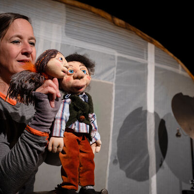 Auf dem Foto ist auf der linken Seite eine weiblich gelesene Person zu sehen, die zwei Handpuppen in den H&auml;nden h&auml;lt. Eine der Handpuppen ist weiblich gelesen, die andere Handpuppe m&auml;nnlich gelesen. Hinter der puppenspielenden Person befindet sich ein wei&szlig;er Stoff, der aus mehreren Teilen zusammengesetzt wurde. En runder, br&auml;unlicher Bogen trennt den wei&szlig;en Stoff von einem schwarzen Hintergrund in der rechten oberen Ecke ab. Vor dem wei&szlig;en Hintergrund steht ganz rechts zudem ein Becken eines Schlagzeuges. Die puppenspielende Person tr&auml;gt ein graues lang&auml;rmliges Oberteils und hat schulterlanges dunkelblondes, glattes Haar.