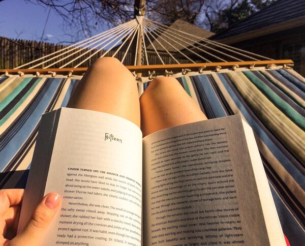 Bild vergr&ouml;&szlig;ern: Perspektive aus der Ich-Sicht: Eine Person liegt in einer gestreiften H&auml;ngematte im Garten und liest ein aufgeschlagenes Taschenbuch, im Hintergrund die eigenen angewinkelten Beine, ein Zaun, B&auml;ume und ein Hausdach vor blauem Himmel.