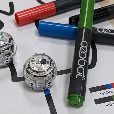 Das Bild zeigt zwei Ozobot-Roboter neben denen farbige Stifte liegen.