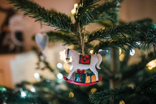 Bild vergr&ouml;&szlig;ern: Wei&szlig;es Schaukelpferd mit kleinen Geschenken als Christbaumschmuck an einem beleuchteten Weihnachtsbaum.