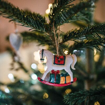 Wei&szlig;es Schaukelpferd mit kleinen Geschenken als Christbaumschmuck an einem beleuchteten Weihnachtsbaum.