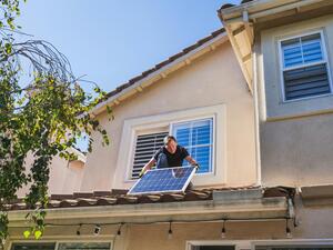 Ein Mann hält eine Photovoltaik-Platte auf dem Vordach seines Hauses in den Händen. 